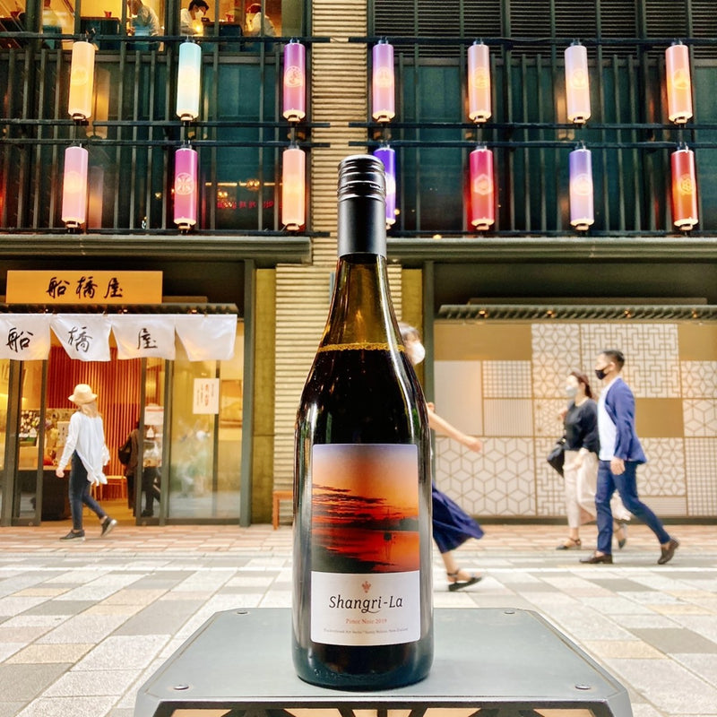 A bottle of Shangri-La Pinot Noir on a little table in the pedestrian zone of an up-market shopping area in Tokyo.  In the background we see people walking past a Japanese delicatessen shop.